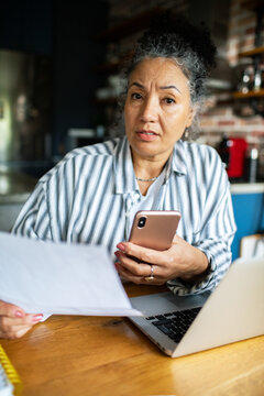 Concerned mature woman reviewing bills with smartphone at kitchen table