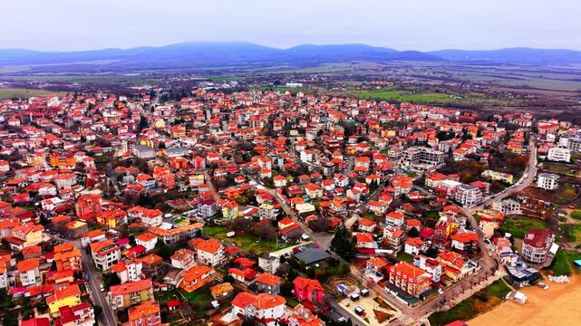 Wide aerial view of Bulgarian town, mountains. Dense red rooftops spread across a valley with distant blue mountain ridges in the background.