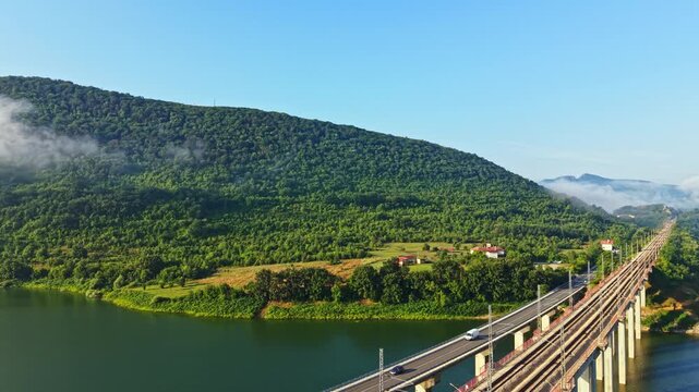 Elevated bridge arcs beside wide river as small vehicles move steadily across span, with calm water below, green hills rising beside route and soft mist under clear blue sky, with bridge, river