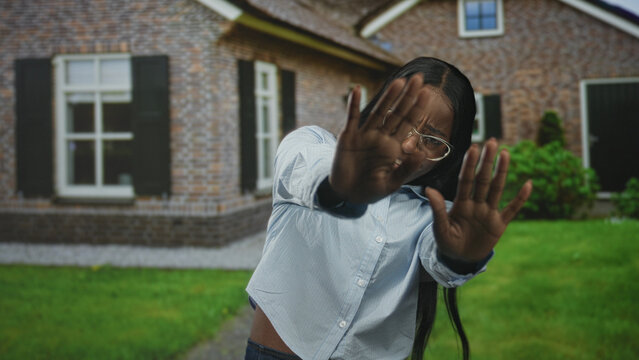 Woman holds up palms to block camera, hands splayed shielding face in front of building with brick facade, window and lawn; privacy.