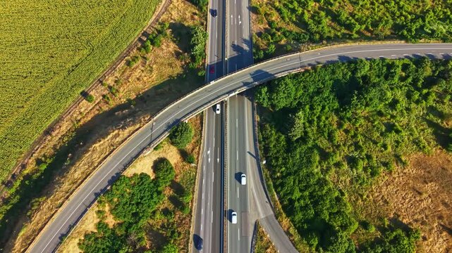 Cars travel along multiple lanes of a highway that intersects fields and trees. The scene shows a mix of asphalt and nature with clear skies.