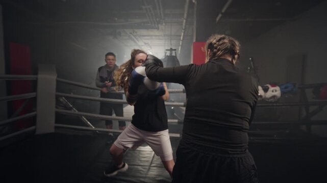 Two young Caucasian women sparring in boxing ring while male coach watches. Clip is suitable for fitness brand commercials and female empowerment campaigns