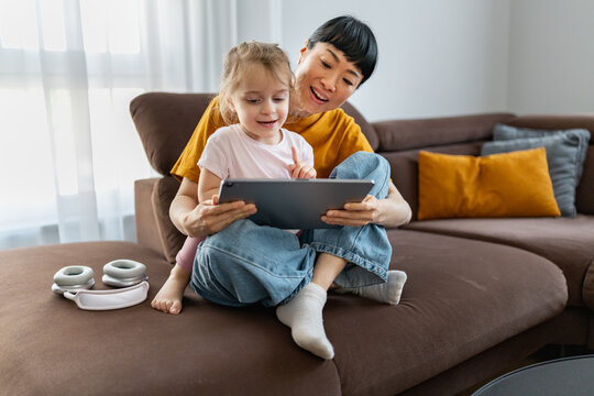 Japanese nanny playing with 3-year-old Caucasian girl on tablet during professional childcare at home