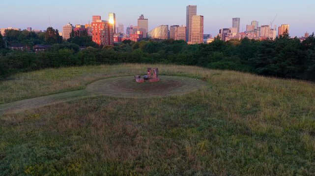 Aerial video overlooking lush green fields at Peachstone Henge towards Victoria Riverside, NOMA, and the glowing sunrise skyline of Manchester city centre.