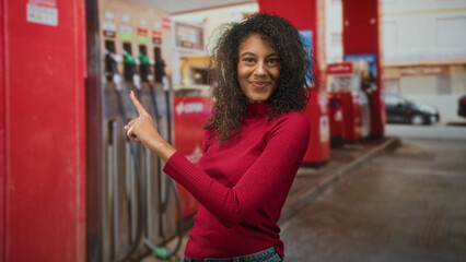 Young woman with curly hair wearing white headphones points finger toward a gas pump at a street station while smiling in a red sweater and denim jeans  joyful curiosity. © Krakenimages.com