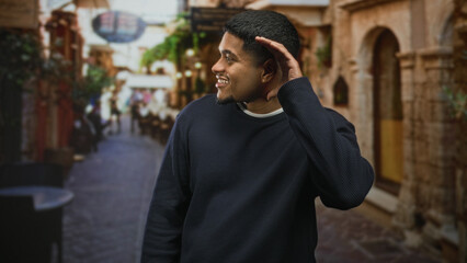 Fototapeta premium Man cupping ear with hand and smiling on a cobblestone street in front of stone buildings; curiosity warmth.