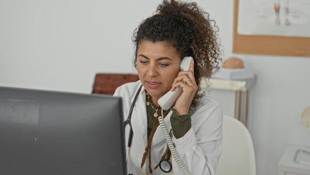 Woman doctor holds telephone to ear while working at computer with stethoscope in clinic building; focused care coordination.