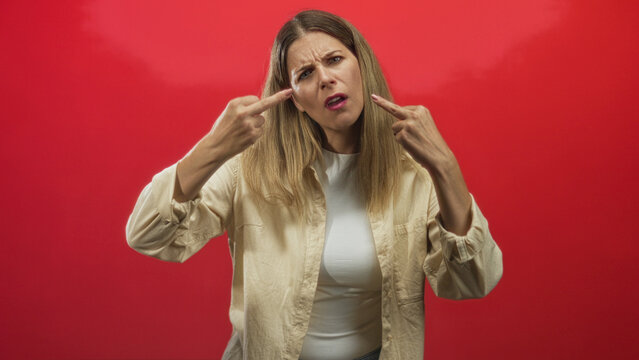 Woman displays raised middle fingers and a stern facial expression toward camera in a red studio backdrop, wearing a beige shirt and white top; defiance rebellion.
