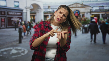 Obraz premium Woman in a red plaid shirt holds hand out with pinched fingers and palm up, asking gesture in a busy street plaza; frustration plea.