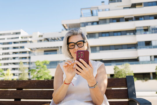 Senior woman using phone on bench near luxury apartments in summer