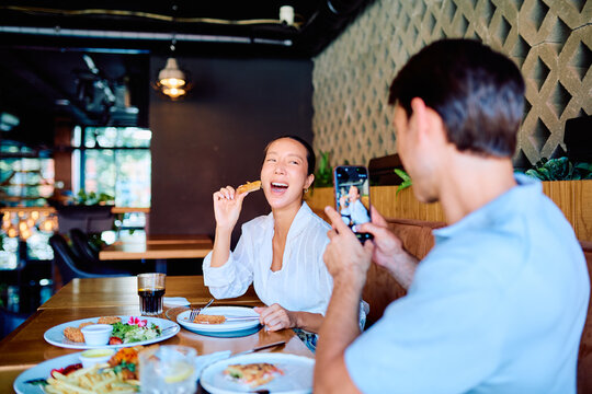 Man taking a smartphone picture of a happy woman eating and laughing at a restaurant, sharing a moment and creating social media content