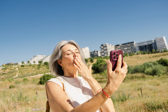 Mature senior woman blowing kiss on video call in summer park blue sky