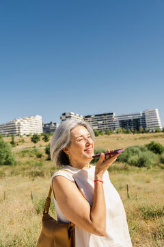 Mature senior woman sending voice message in summer park copyspace