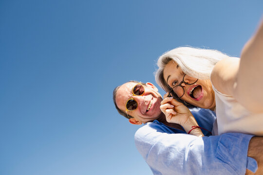 Playful senior couple taking low angle selfie under blue sky