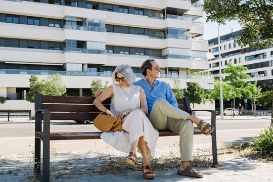 Senior couple sitting on bench near luxury apartments in summer