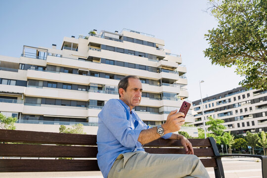 Senior man checking phone on bench by luxury apartments in summer
