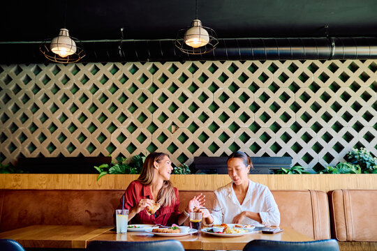 Two diverse women friends sitting at a table in a modern restaurant, eating food, drinking, and having a lively conversation