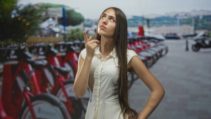 Fototapeta premium Young woman finger to chin thoughtful gesture by row of red rental bikes at street bike station; confidence.