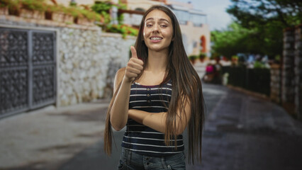 Fototapeta premium Teenage girl with braces smiles and gives thumbs up with arm crossed, wearing a striped tank top and jeans on a sunny street; confidence.