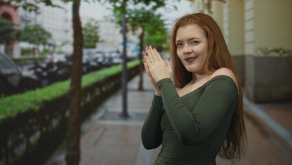 Fototapeta premium Redhead woman clapping hands on city street sidewalk with exposed shoulder and green off shoulder top; joyful confidence.