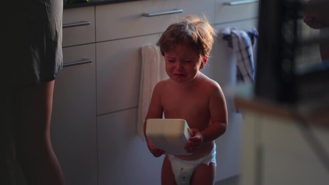 Crying toddler holding food container in kitchen, expressing frustration, unmet needs and emotional overwhelm