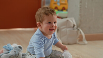 Fototapeta premium Boy holding wooden toy and white cloth with hand while sitting on floor in building near plush rabbit and diaper bag; joyful play.