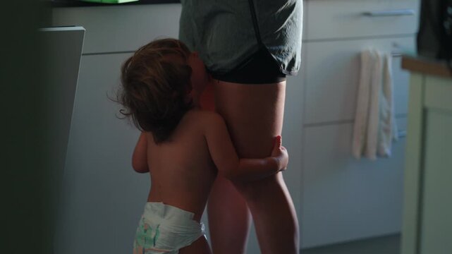Crying toddler hugging parent&rsquo;s leg in kitchen, seeking comfort, reassurance and emotional security