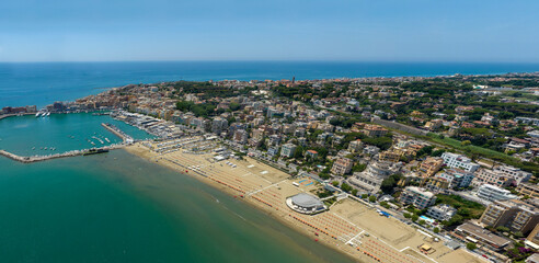 Fototapeta premium Aerial view of Anzio, in Lazio, Italy. It's a coastal city featuring a busy harbor, sandy beaches, and urban buildings along the shore under a clear sky, capturing an ideal summer destination.