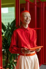 Fototapeta premium Smiling redheaded professional in bright red shirt holding a tablet in a modern red office interior for corporate portrait and business branding materials