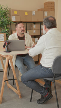 Two men talking over a laptop beside stacked cardboard packages, one leaning on the table and pointing at labels in a building; collaboration.