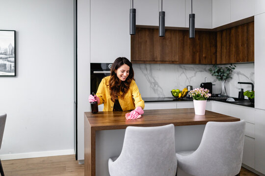 Woman cleaning modern kitchen island with spray bottle and pink gloves, smiling while tidying