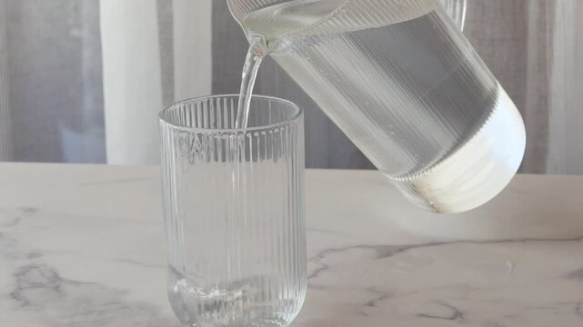 Fresh drinking water pouring from modern glass pitcher into textured glass for healthy hydration.