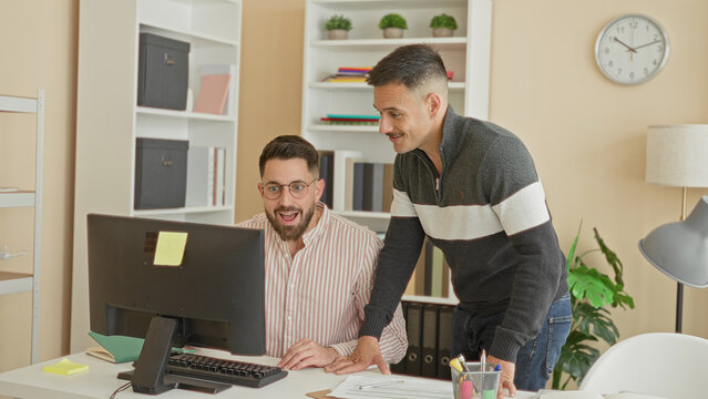 Two men at a computer, seated worker raising hand for fistbump while standing colleague leans over the desk in building; teamwork camaraderie.