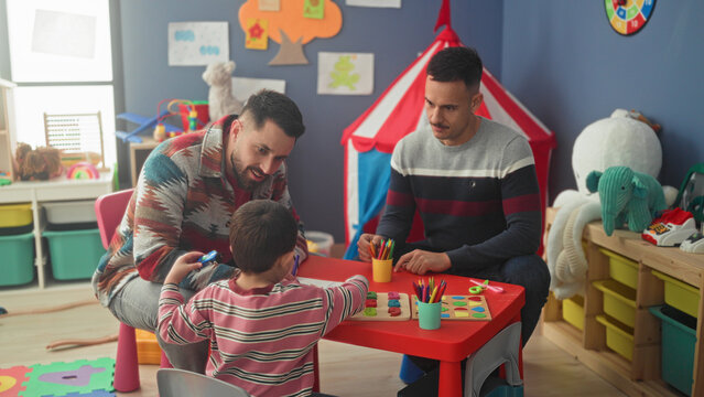 Boy sits at small red table playing with crayons and wooden puzzle while two men point and guide with hands in a building; playfulness bonding.
