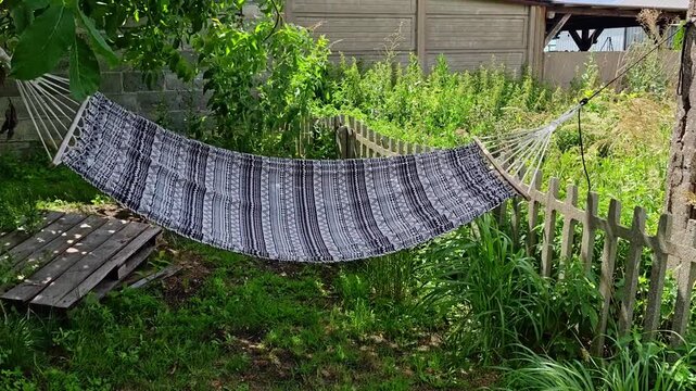 Swinging hammock in the wind with black and white ethnic pattern stretched in a rustic garden between a tree and fence, lush green grass around, wooden pallet, natural sunlight.