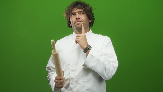 Man chef in white uniform holding a wooden rolling pin, finger to chin and eyes closed against green backdrop in studio; thoughtful deliberation.