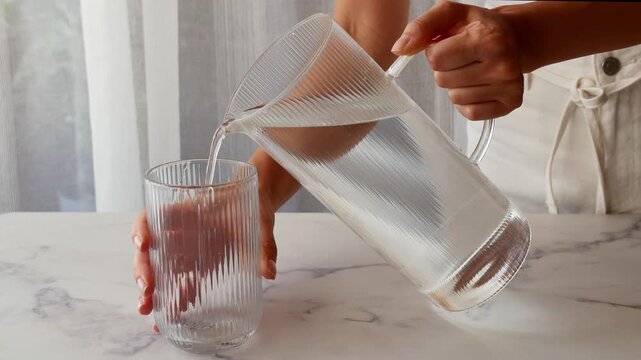 Woman pouring fresh drinking water from glass pitcher into textured glass for healthy hydration