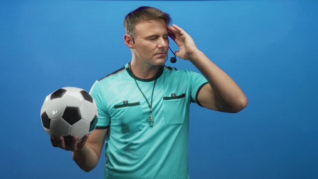 Man holding a soccer ball in one hand and touching his temple with the other hand, referee with whistle and headset visible in studio; focus strategy.