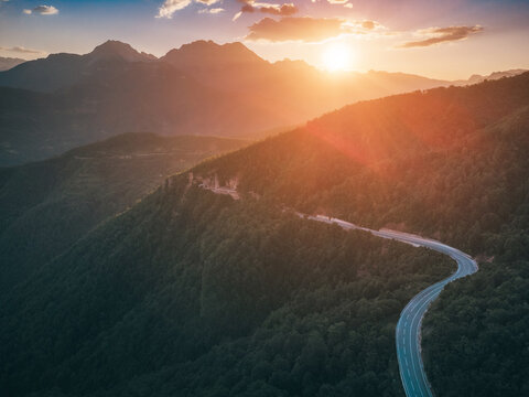 Winding mountain road traversing a lush green forest with dramatic sun flare and mountain peaks during a vibrant sunset