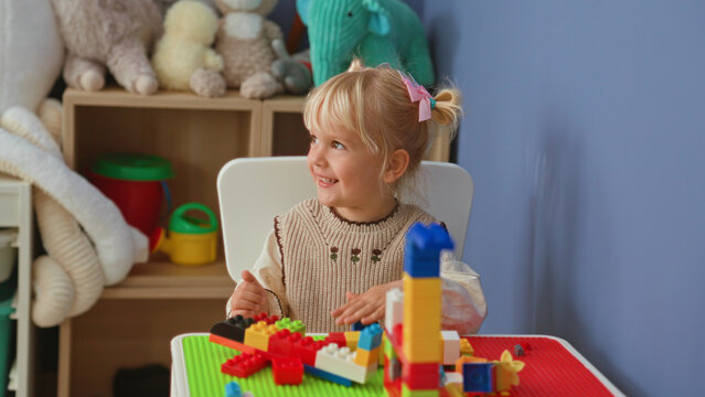 Little blonde girl building a colorful block tower while seated at a playroom table with stuffed plush toys on a shelf nearby; playful curiosity.