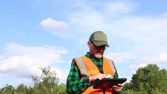 Ecologist, climatologist or environmental supervisor in protective uniform analysing weather data on a digital tablet in front of a green forest and blue sky.	