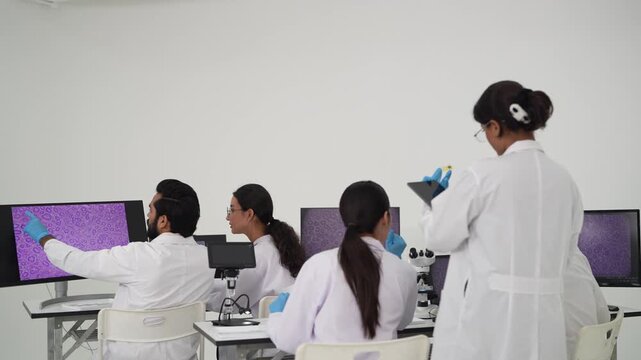 Row of seated laboratory scientists in white coats reviews magnified tissue images across multiple monitors during a focused pathology analysis session at work