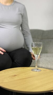 Expecting mother holding a glass of wine on sofa, showing unhealthy pregnancy habits.