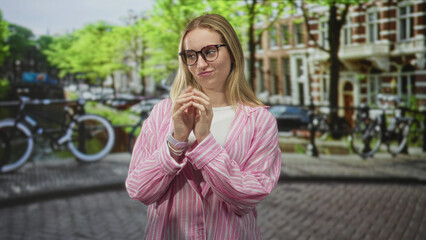 Fototapeta premium Woman clasping hands, pink striped shirt and glasses on a canal street with bicycles and brick buildings; thoughtful reflection.