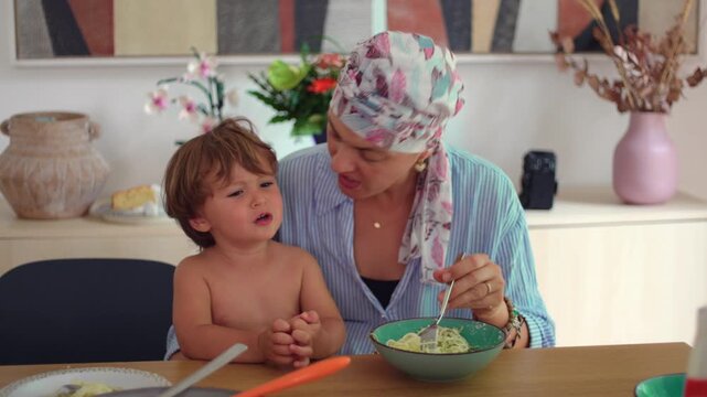 Mother with cancer feeding pasta to her upset toddler at the dining table at home, nurturing family connection expressing resilience, comfort and parental devotion
