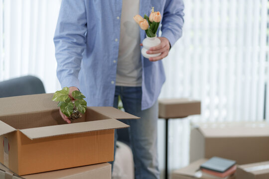 Asian man unpacking cardboard box holding potted plant and flowers while settling into new home