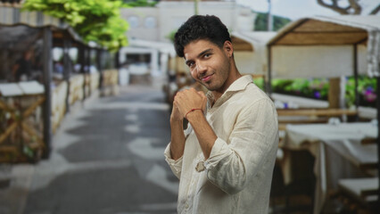Fototapeta premium Man in a light linen shirt smiles with clasped hands near chest at a busy street cafe market row, facing camera; joyful relaxed confidence.