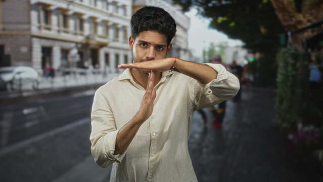 Man with bare hands making a time out gesture on a busy street by building facades, wearing beige linen shirt and looking directly at camera; confidence.