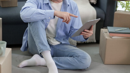 Asian man pointing at tablet screen while sitting on floor surrounded by moving boxes in new apartment © snowing12
