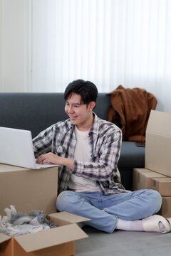 Young Asian man using laptop while sitting on floor surrounded by cardboard boxes during home relocation